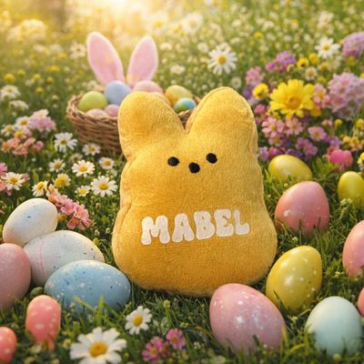 Soft yellow Easter bunny plush toy personalised with the name Mabel, sitting on grass amongst pastel Easter eggs and daisies 
