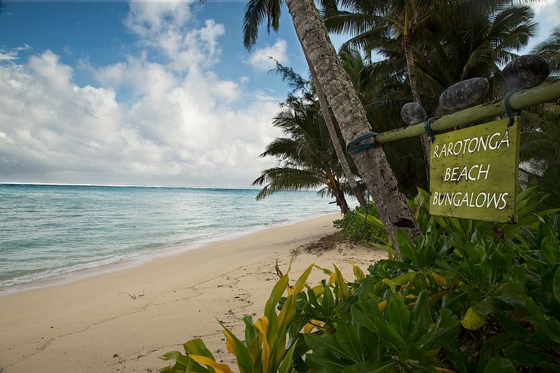 Sign saying "Rarotonga Beach Bungalows" overlooking the beach with palm trees.