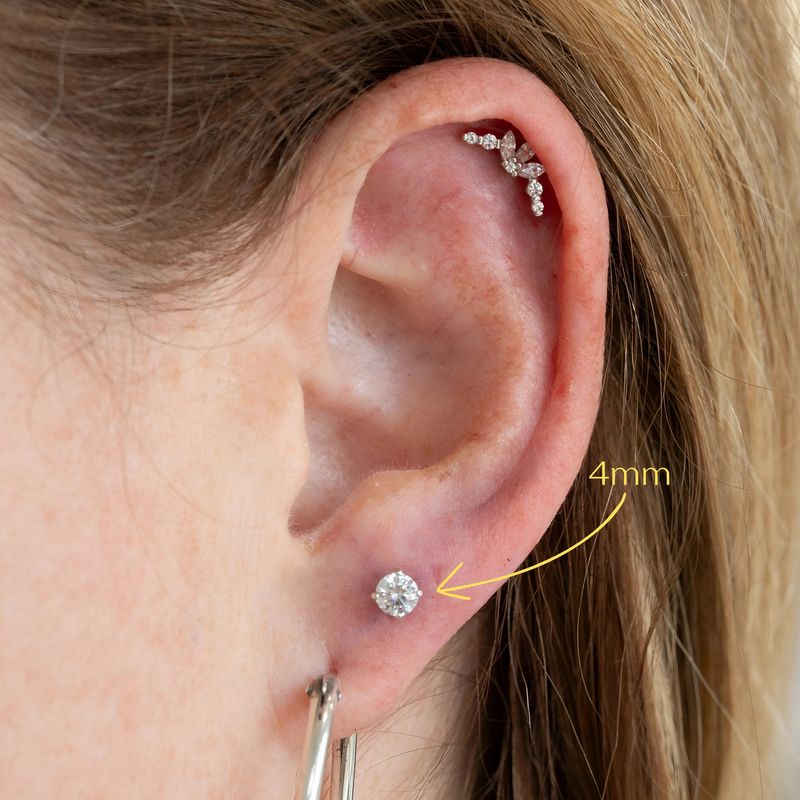 Close-up of an ear with piercings. A 4mm round cubic zirconia stud is inserted in the second earlobe placement, highlighted with a yellow arrow and text that says “4mm”. The upper ear has a helix piercing with a decorative, curved piece with multiple small gemstones and a hoop earring is worn in the first lower lobe piercing.