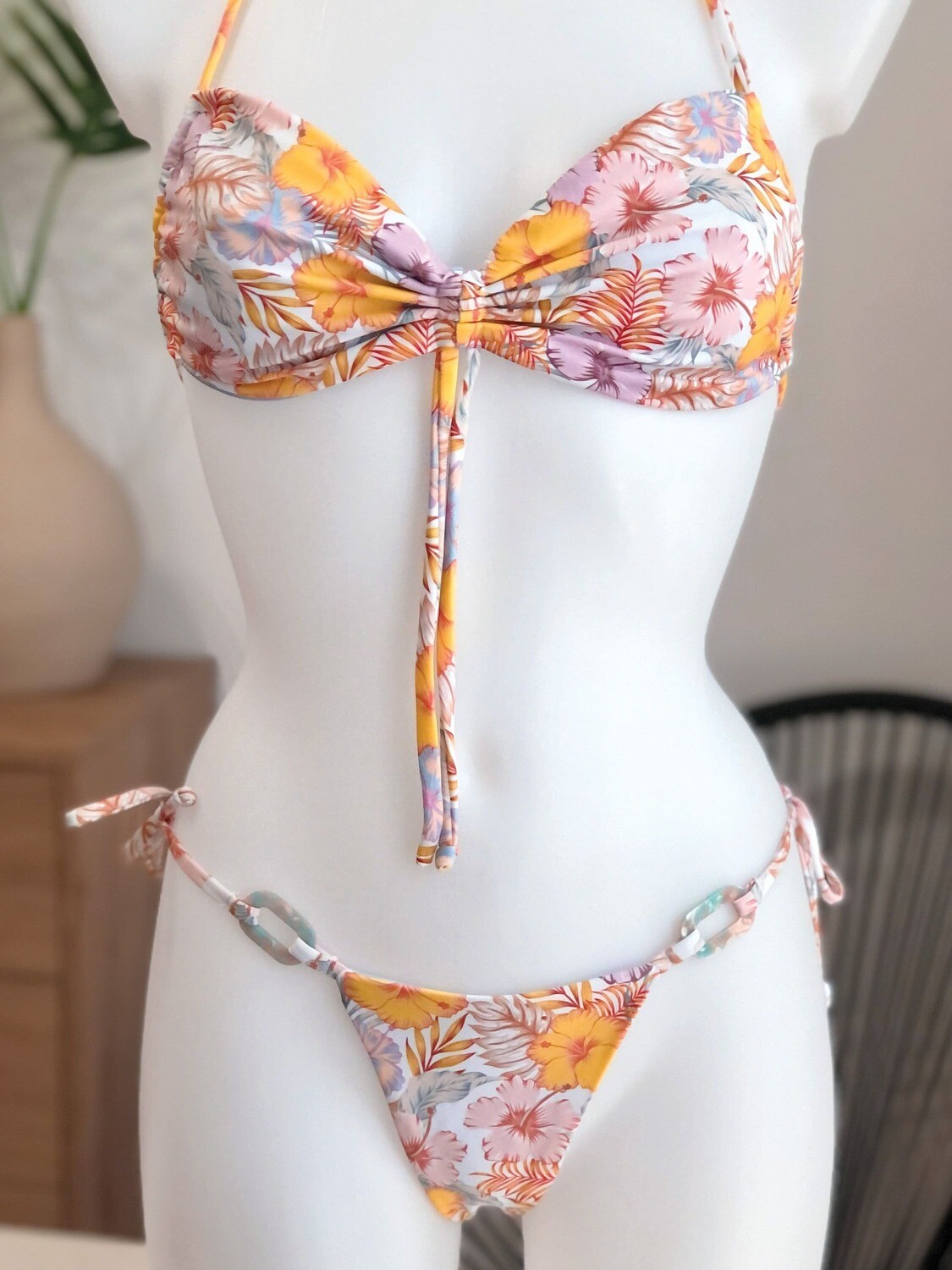 Floral bikini with pastel tones featuring a ruched bandeau front-tie bikini top and matching tie tanga bottoms. The tropical print in soft pink, yellow, and purple hues creates a delicate and feminine beach look. Made with eco-friendly, high-end fabric, perfect for stylish and sustainable summer wear.
