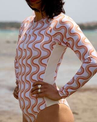 Woman wearing a long-sleeve one-piece swimsuit with a cream and terracotta wavy retro-chic pattern, standing on a beach with an ocean backdrop. The swimsuit features a front zipper and comfortable design, perfect for sun protection and water activities.