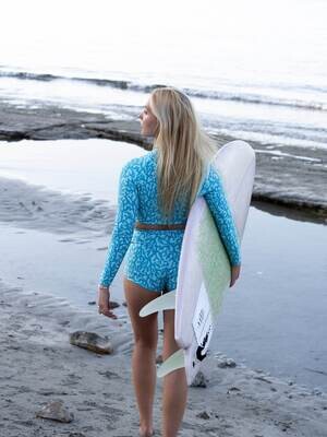 Woman at the beach holding a surfboard in a turquoise coral print high-waist retro bottom shorts and eco-friendly long-sleeve crop top rash guard.