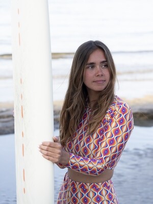 Woman holding a surfboard in eco-friendly long-sleeve crop top rash guard and high-waist retro bottom shorts with magenta, purple, orange, and blue symmetrical retro print.
