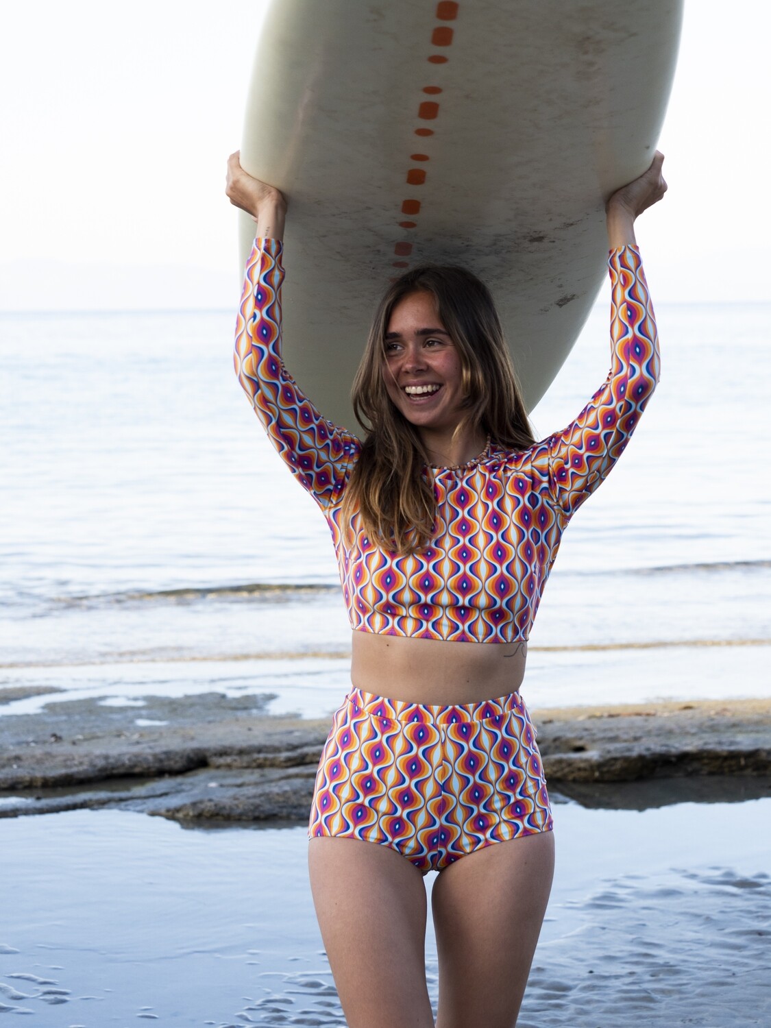 Eco-friendly long-sleeve crop top rash guard and high-waist retro shorts in magenta, purple, orange, and blue print worn by a woman at the beach with a surfboard.