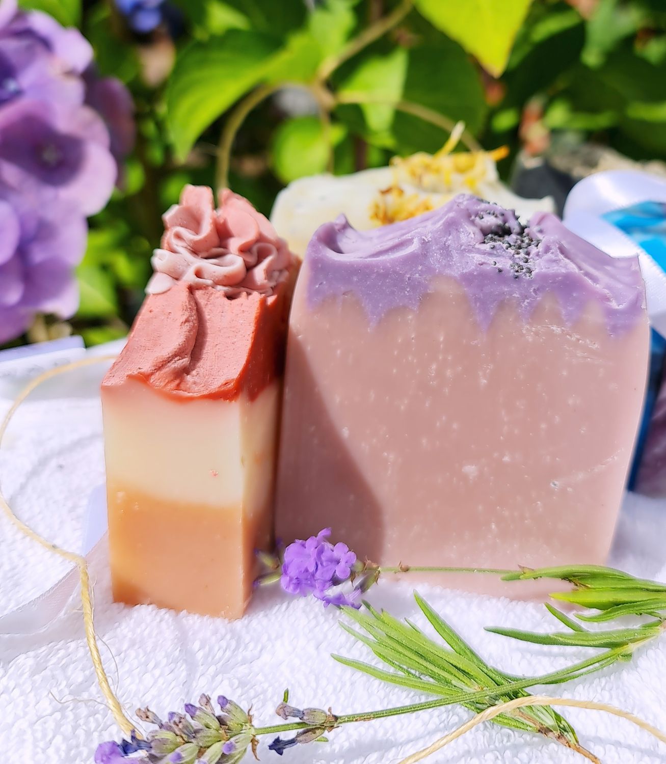 Handcrafted lavender soap bars with natural ingredients, displayed on a white towel surrounded by fresh lavender and garden flowers. One soap features a pink and cream swirl, the other a purple lavender design, highlighting their eco-friendly, vegan, and cruelty-free qualities. Perfect for natural skincare enthusiasts.&quot;