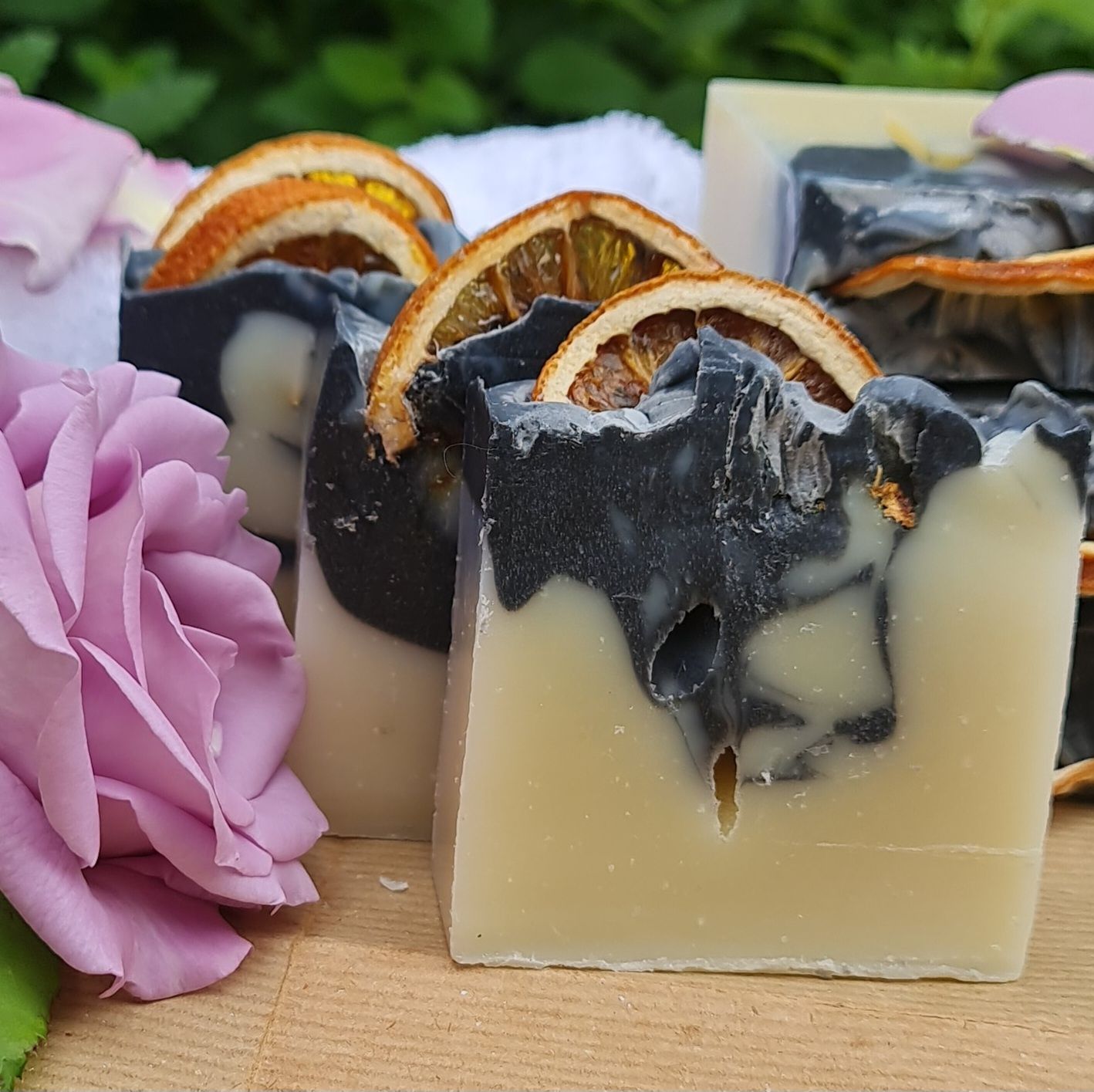 Handcrafted soap bars with a black and cream layered design, topped with natural dried orange slices, displayed beside a pink rose for a luxurious and natural skincare presentation