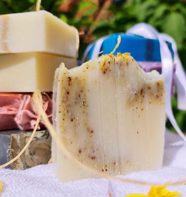 Handcrafted cold process soap bar with natural exfoliants, set against a background of stacked soap bars in various colors, displayed on a white towel with green foliage in the background