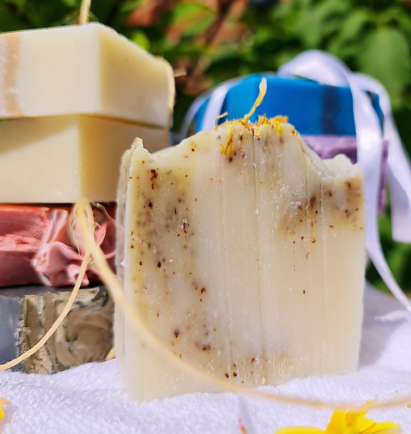 Handcrafted cold process soap bar with natural exfoliants, set against a background of stacked soap bars in various colors, displayed on a white towel with green foliage in the background