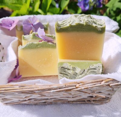 Handcrafted soap bars with a green and yellow layered design, placed in a wicker basket lined with a white towel, accented by purple flowers in a garden setting.