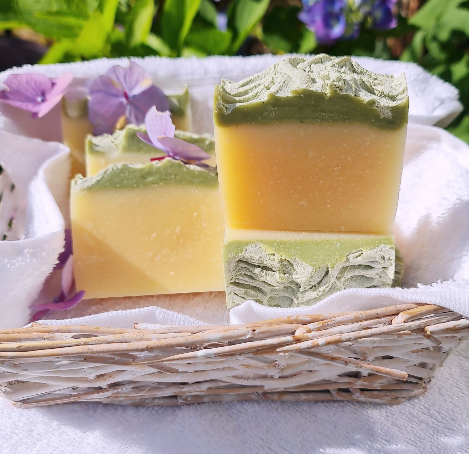 Handcrafted soap bars with a green and yellow layered design, placed in a wicker basket lined with a white towel, accented by purple flowers in a garden setting.