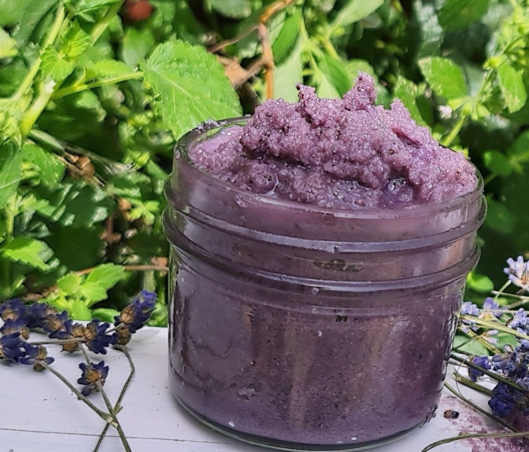 Handcrafted lavender sugar scrub in a glass jar, displayed outdoors with fresh lavender flowers and green foliage. This natural exfoliating scrub is vegan, cruelty-free, and made with essential oils and organic ingredients, perfect for a luxurious and eco-friendly skincare routine