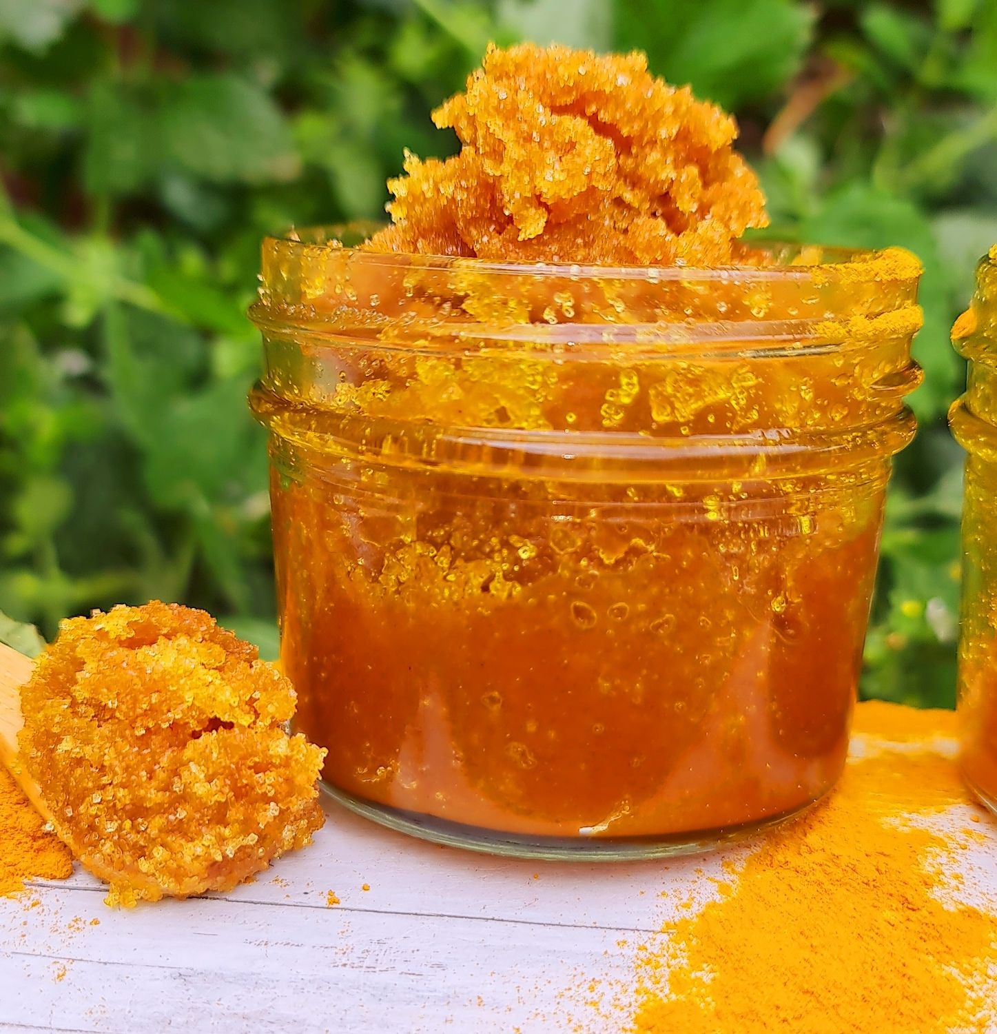 Turmeric &amp; Lemon Sugar Body Scrub in a clear glass jar, showcasing the vibrant golden color and coarse texture of the natural exfoliating scrub, with a backdrop of greenery emphasizing the natural and organic ingredients