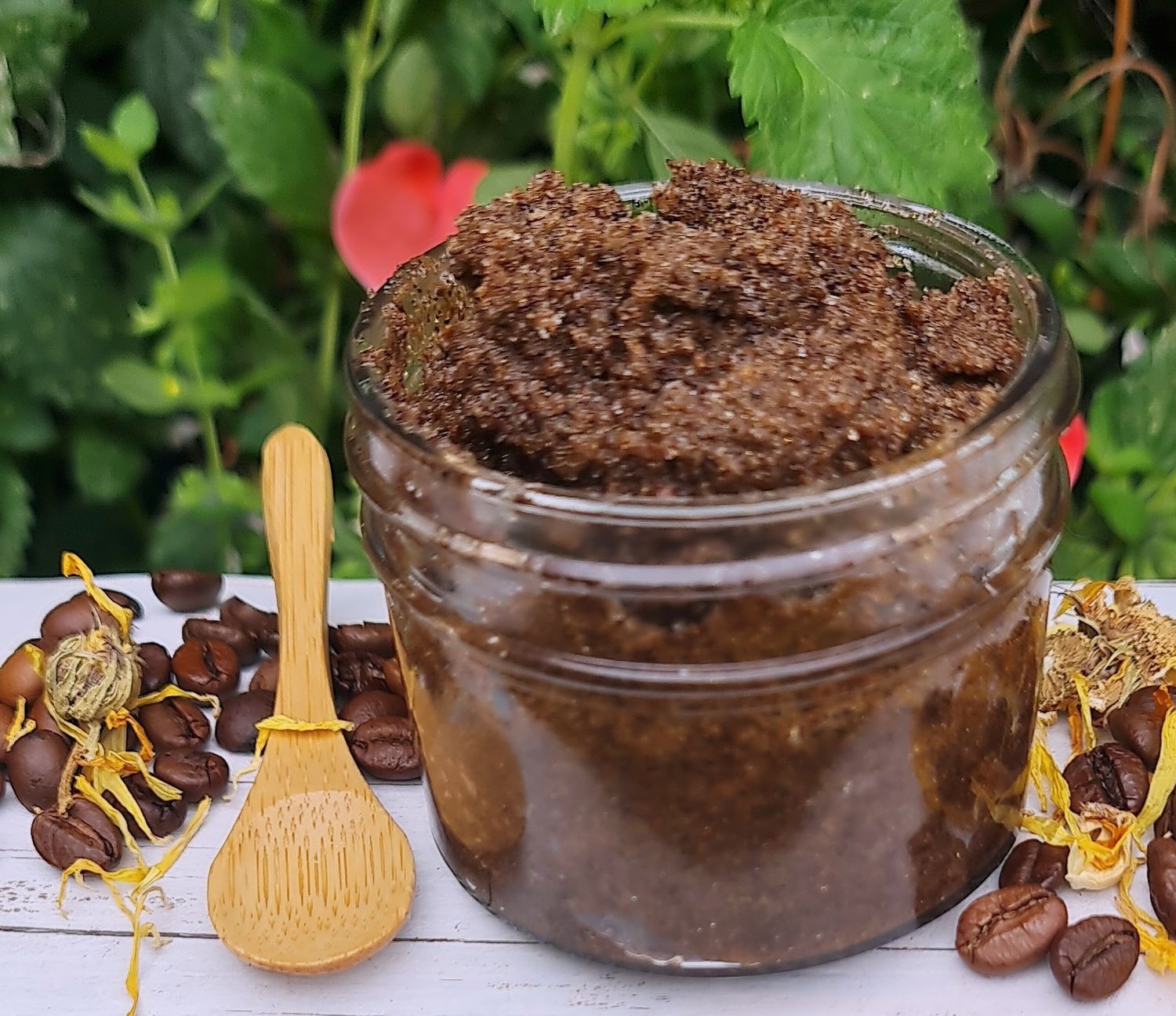 Jar of natural coffee scrub surrounded by coffee beans, dried flowers, and a small wooden spoon, displayed on a rustic wooden surface with green foliage in the background.&quot;