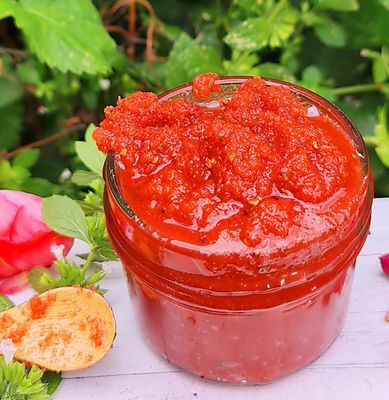 Bright red Raspberry Seeds Sugar Scrub in a glass jar, surrounded by fresh green leaves and pink flowers, showcasing the vibrant, natural exfoliating skincare product.