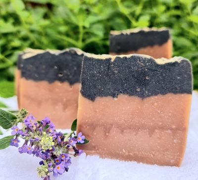 Tobacco &amp; Vanilla handcrafted cold process soap bars with a dual-tone design, featuring a black activated charcoal top and a natural beige bottom, set against a green leafy background with a sprig of purple flowers in the foreground.