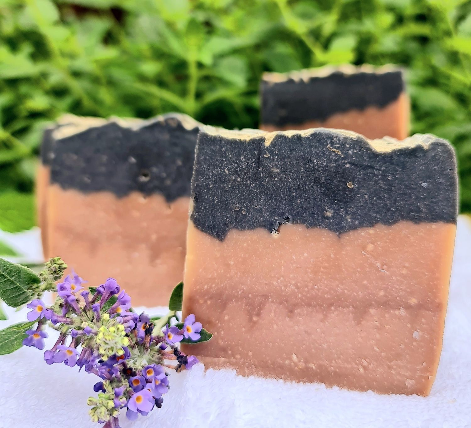 Tobacco &amp; Vanilla handcrafted cold process soap bars with a dual-tone design, featuring a black activated charcoal top and a natural beige bottom, set against a green leafy background with a sprig of purple flowers in the foreground.