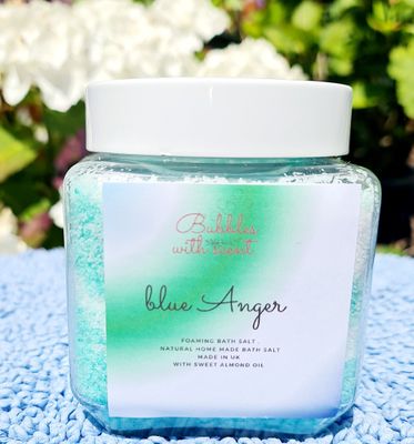 Jar of Blue Anger foaming bath salts by Bubbles with Scent, featuring a green and white label, placed on a blue textured surface with a garden background.