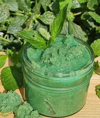 Jar of green Peppermint Sugar Scrub on a wooden surface, topped with fresh mint leaves, surrounded by green foliage in the background.