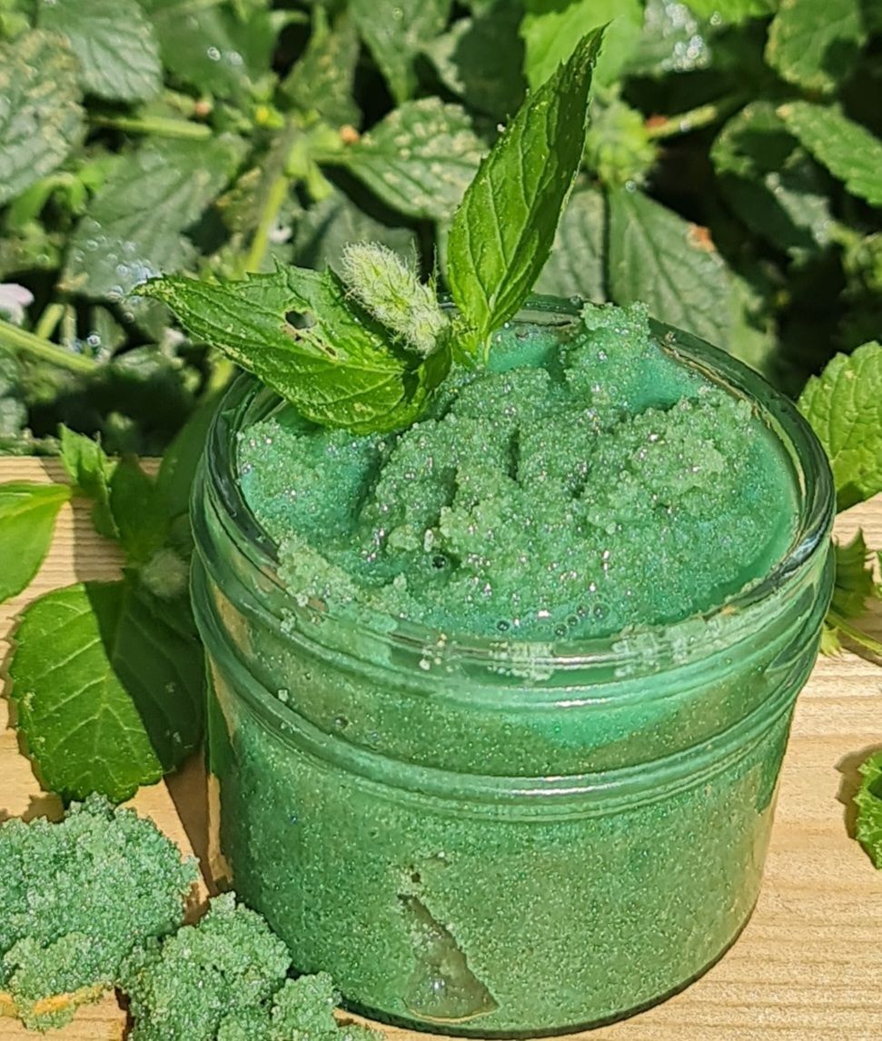 Jar of green Peppermint Sugar Scrub on a wooden surface, topped with fresh mint leaves, surrounded by green foliage in the background.