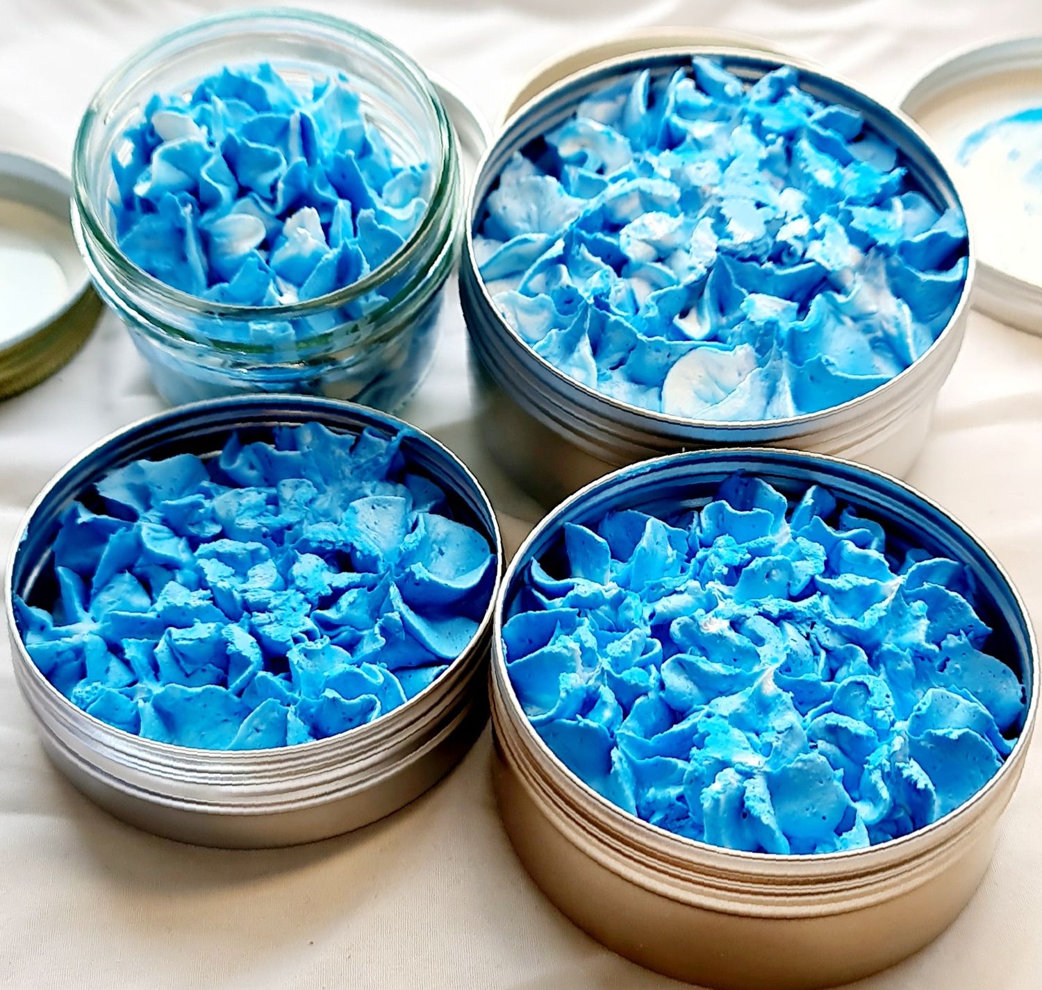 Four containers of All Smells Better in Blue whipped soap in eco-friendly packaging. The soap has a vibrant blue, fluffy, and creamy texture, displayed in both glass and metal tins.