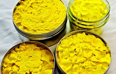 Four containers of All Smells Better in Yellow whipped soap, showcasing a vibrant yellow, fluffy texture. The soap is packaged in eco-friendly glass jars and metal tins, placed on a white surface.