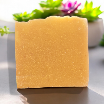 A single handmade bar of coconut and turmeric soap displayed upright on a surface with a blurred background of green and purple plants