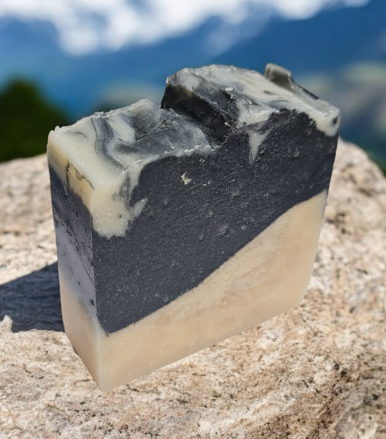  A handmade bar of natural soap with black and beige layers, featuring activated charcoal, placed on a rock surface with a blurred mountain landscape in the background