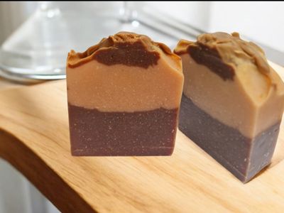 Two bars of layered soap with rich brown and tan shades, displayed on a wooden surface. The soap bars have a textured top and smooth layers, with a darker bottom half and a lighter top half, showcasing a marbled effect between the layers