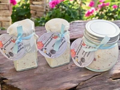 Three jars of Magnesium &amp; Goat Milk Botanical Spa Soak, elegantly packaged with a light blue ribbon and floral tags, displayed outdoors on a rustic wooden surface with a vibrant garden in the background.