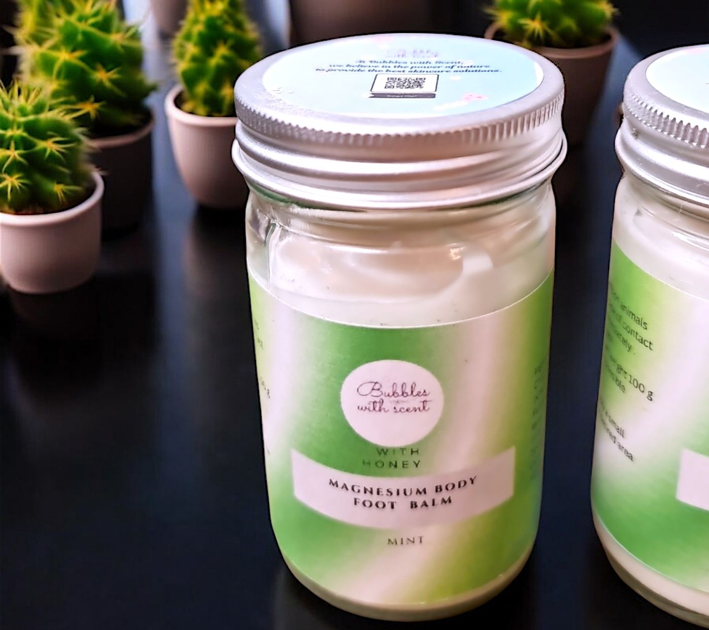 Bubbles with Scent Magnesium Body Foot Balm in a glass jar with a green gradient label, featuring the scent &#39;Mint with Honey.&#39; The jar is placed on a dark surface, surrounded by small potted cacti, highlighting the product&#39;s fresh and natural appeal