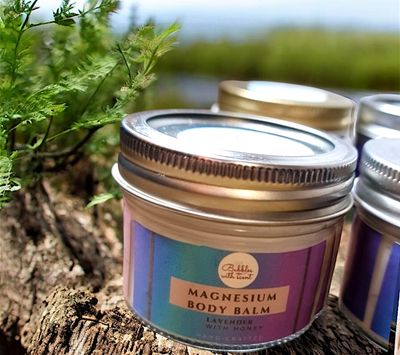 A small jar of Bubbles with Scent Magnesium Body Balm in Lavender &amp; Honey scent, placed outdoors on a rustic wooden surface with greenery in the background. The jar features a purple gradient label with a calming aesthetic, highlighting the product’s natural and soothing qualities