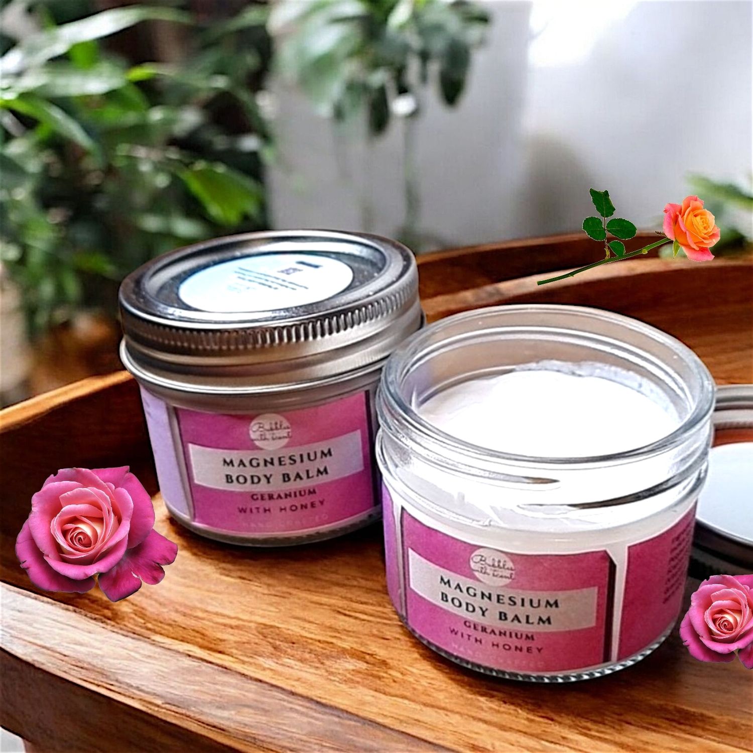 Two jars of Bubbles with Scent Magnesium Body Balm in Geranium with Honey scent, one open and one closed, placed on a wooden tray. The jars have a pink gradient label, complementing the floral theme. Roses and green plants in the background add a natural and calming touch to the setting