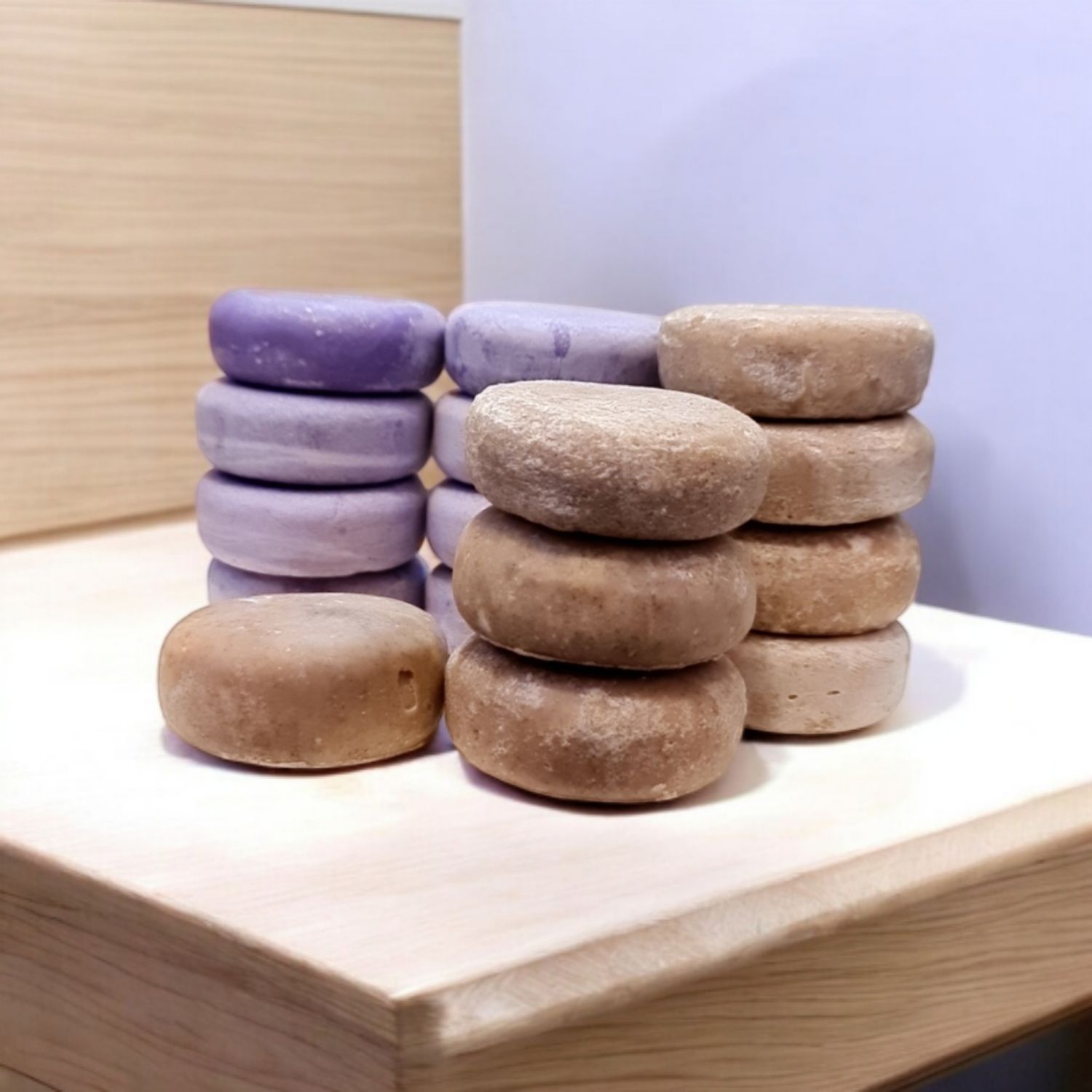 Stacks of handmade round soap bars displayed on a light wooden surface. The soaps are arranged in two colors: a light purple (possibly lavender-scented) and a natural beige tone, highlighting their artisanal quality and natural ingredients