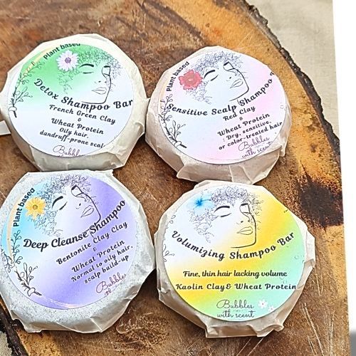 Four round, wrapped shampoo bars from Bubbles with Scent, displayed on a rustic wooden surface. Each shampoo bar has a distinct label with a watercolor background and an illustration of a woman&#39;s face with floral accents. The labels indicate different formulations:

&#39;Detox Shampoo Bar&#39; – French Green Clay for oily, dull, dandruff-prone scalp (green-themed label).
&#39;Sensitive Scalp Shampoo Bar&#39; – Red Clay for dry, sensitive, or color-treated hair (purple-pink-themed label).
&#39;Deep Cleanse Shampoo Bar&#39; – Bentonite Clay with Wheat Protein for normal to oily scalp (blue-themed label).
&#39;Volumizing Shampoo Bar&#39; – Kaolin Clay &amp; Wheat Protein for fine, thin hair lacking volume (yellow-green-themed label).