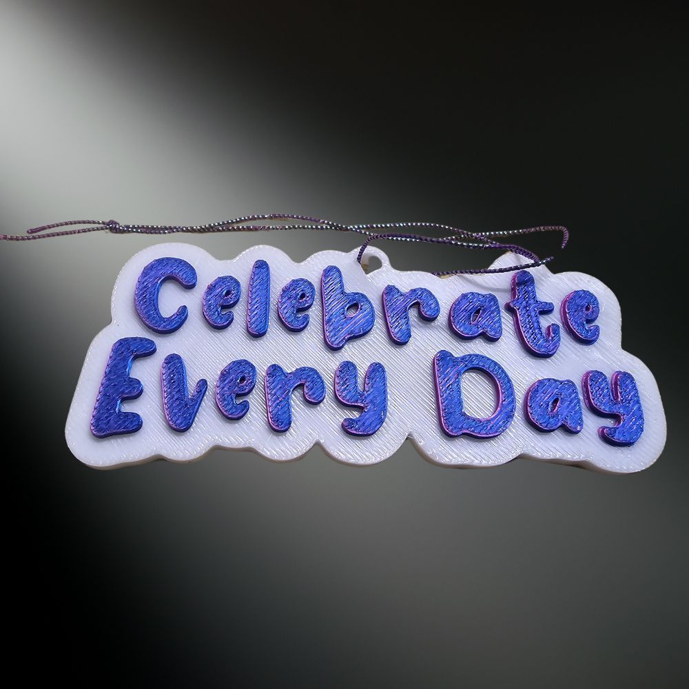 Close-up of the Celebrate Every Day rearview mirror hanger or ornament, featuring purple
 3D lettering on a white background.