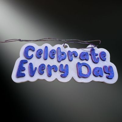 Close-up of the Celebrate Every Day rearview mirror hanger or ornament, featuring purple
 3D lettering on a white background.