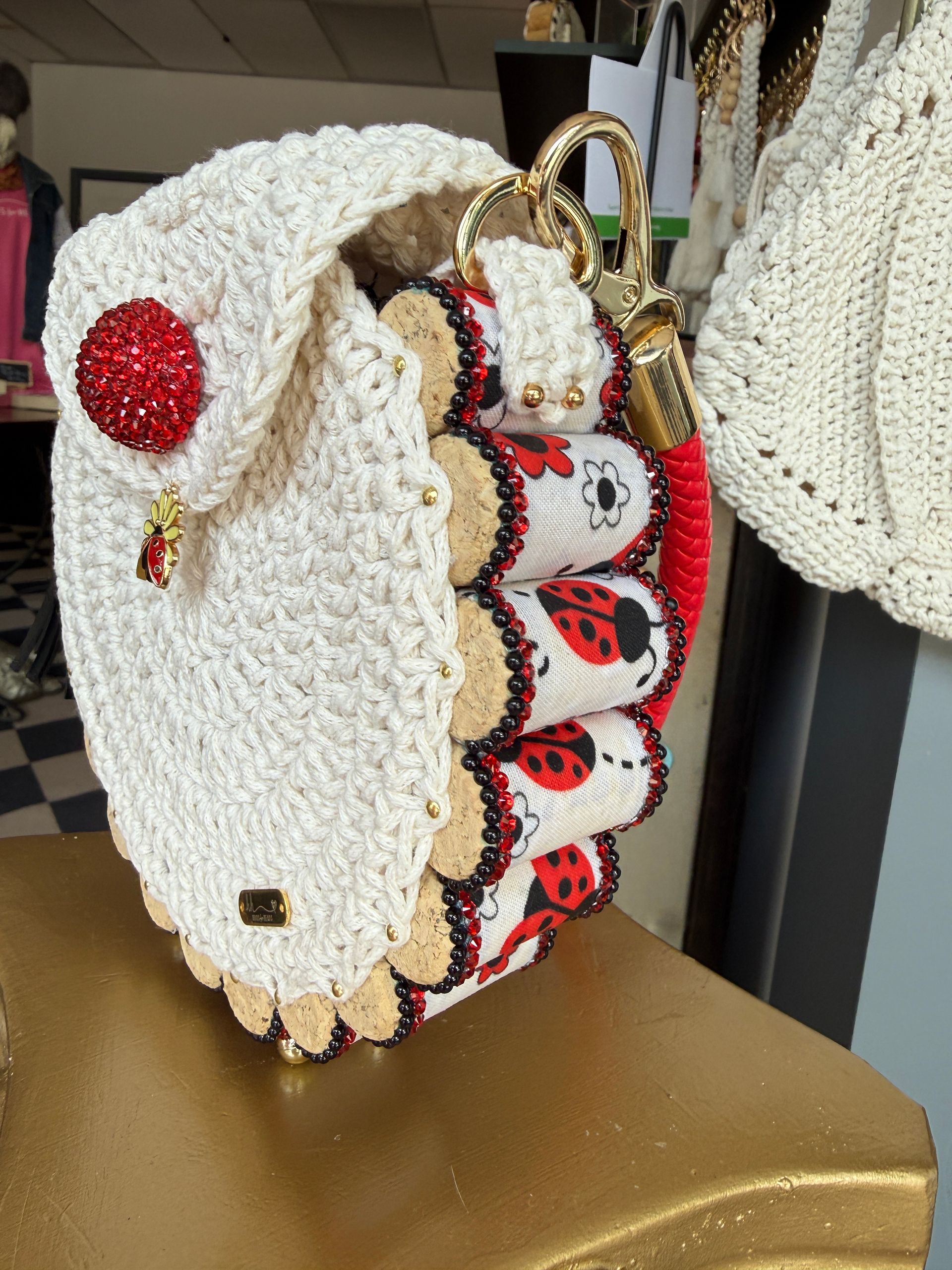 Full view of a handmade cream round crochet bag hanging by a red braided strap, featuring a ladybug charm and scalloped cork