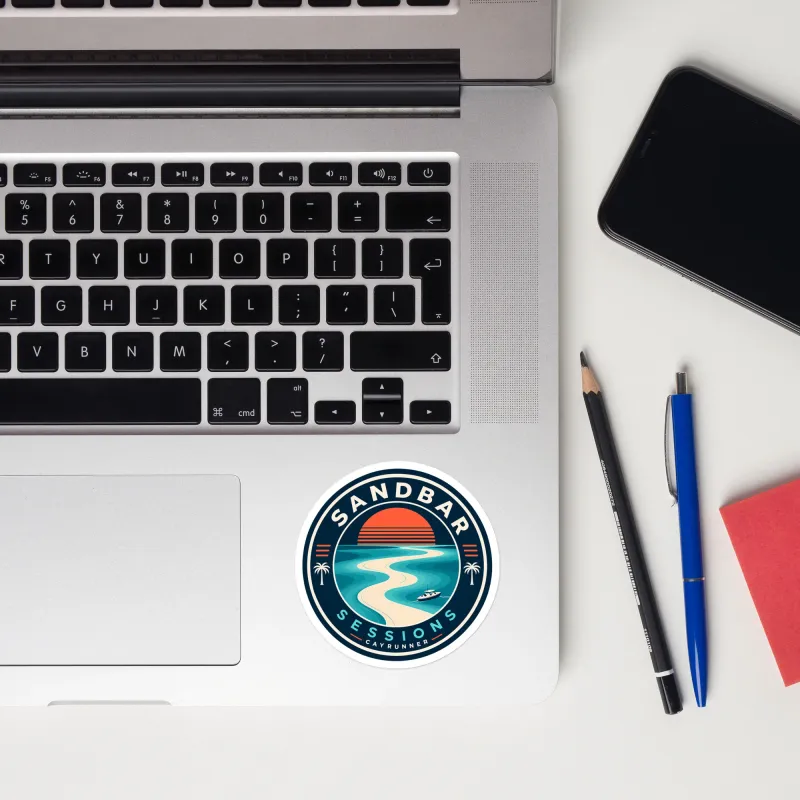 Cayrunner Sunset Sandbar sticker placed on a laptop trackpad area, near keyboard, phone, pens, and a red notepad.