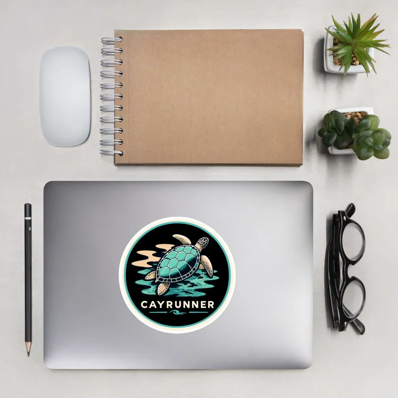 Cayrunner Turtle Drift sticker on a laptop lid, flanked by a mouse, notebook, glasses, pencil, and plants on a desk.