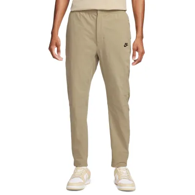 Front view of Nike Commuter woven pants in khaki with tapered legs, open hems and black Nike logo.