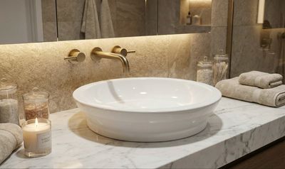 Lavabo de salle de bain en céramique blanche avec robinet doré sur un comptoir en marbre, avec serviettes et bougie à proximité.