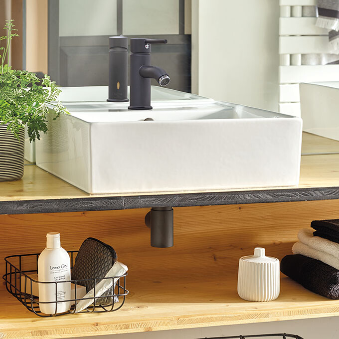 Mini siphon de lavabo Wirquin Linéis noir mat sous lavabo blanc sur étagère de salle de bain en bois.