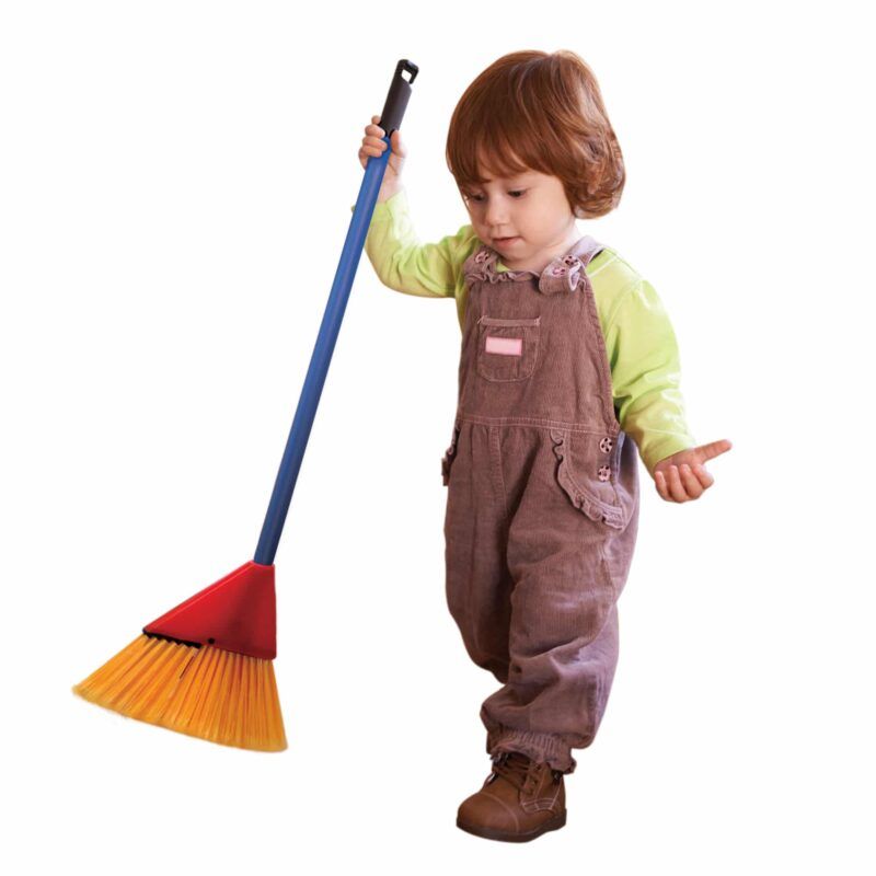 A toddler in brown overalls and green shirt holds a blue broom, looking curious.