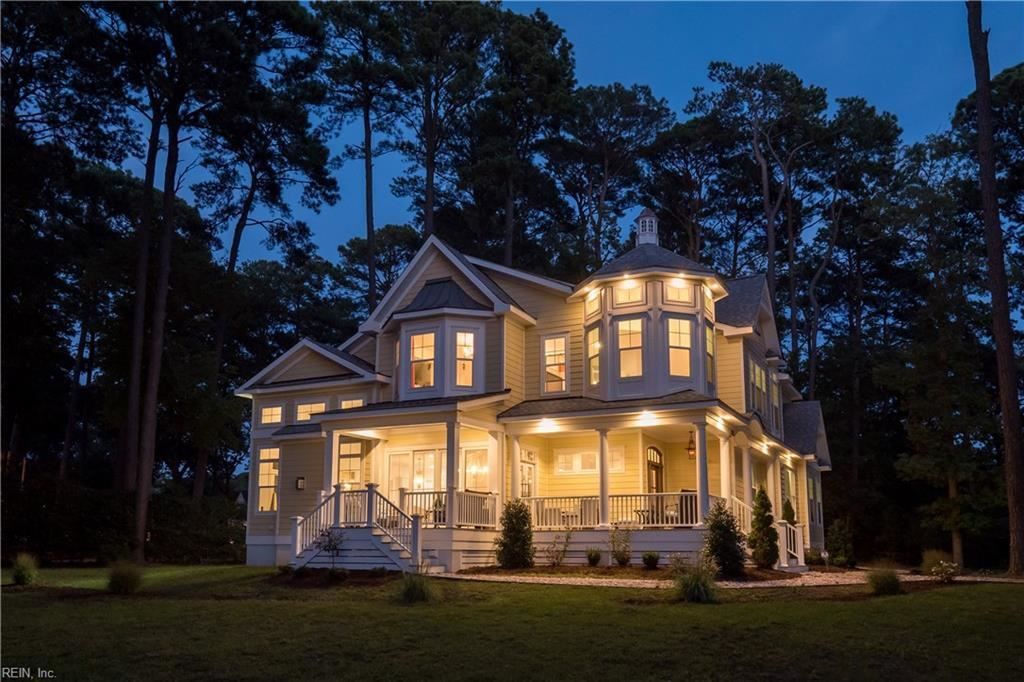 Evening exterior of a large two-story house with warm interior lighting, wraparound porch, and wooded surroundings.
