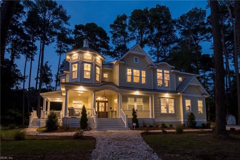 Evening exterior of a large two-story house with warm interior lighting, wraparound porch, and wooded surroundings.
