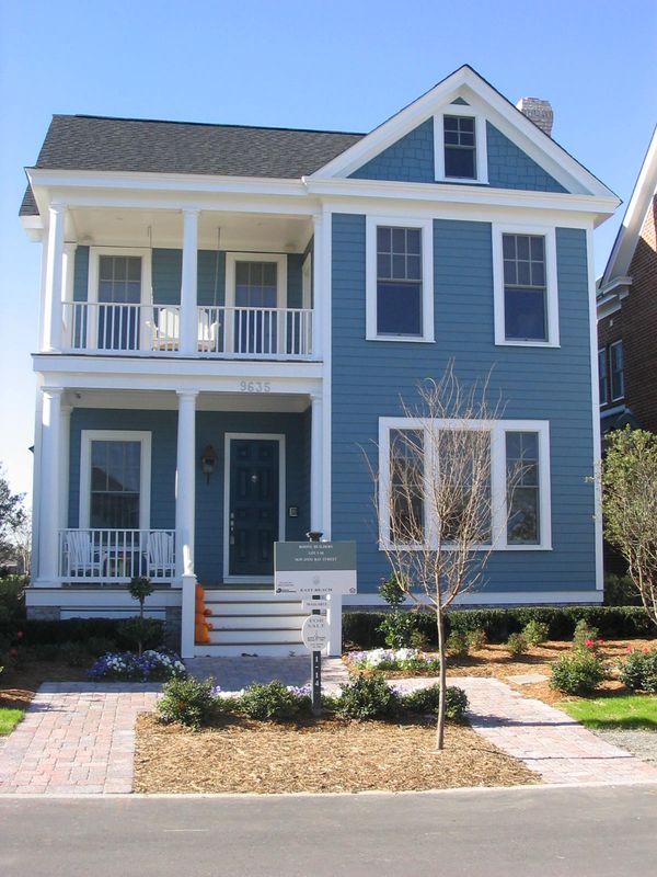 Front exterior of a two-story blue house with white trim, covered porch, balcony railing, and landscaped yard.