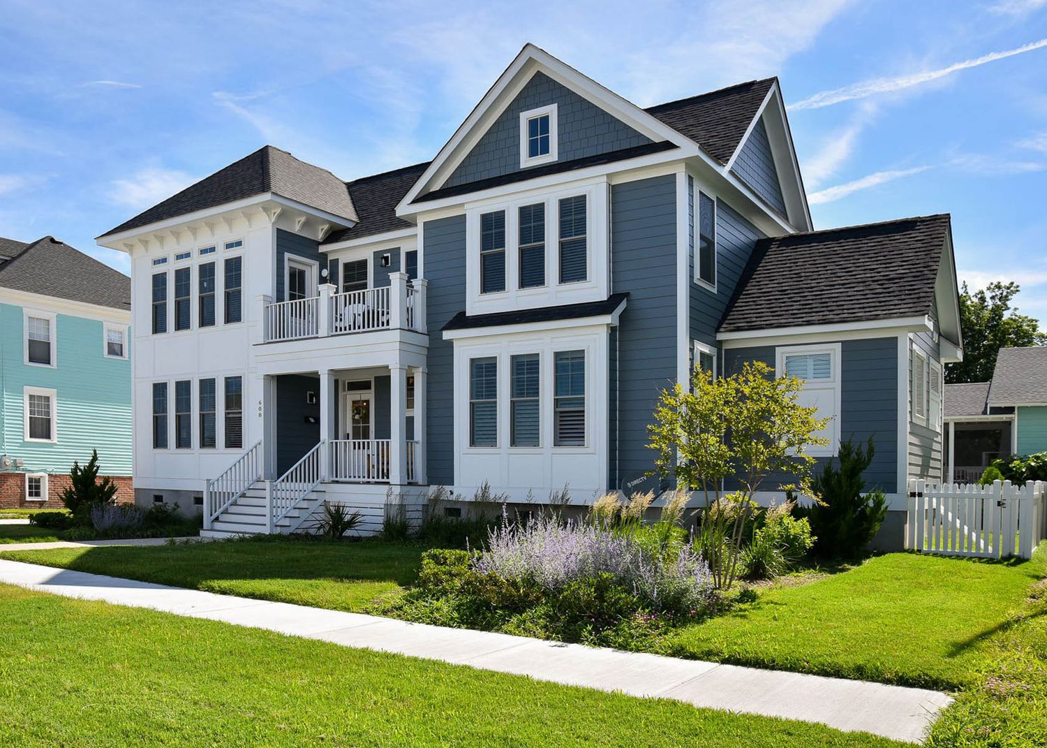 Front view of a two-story blue and white house with gable roof, balcony, lawn, and white picket fence.