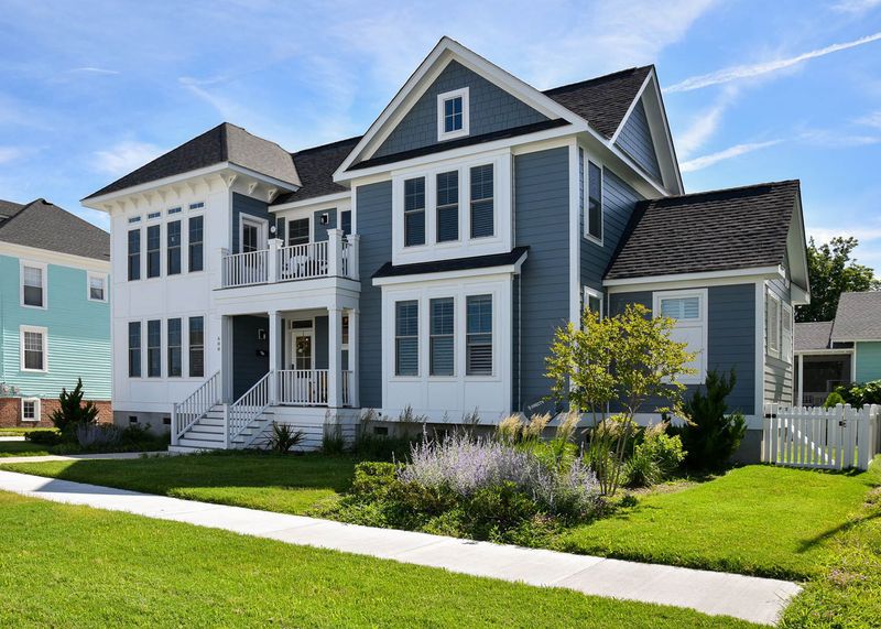 Front view of a two-story blue and white house with gable roof, balcony, lawn, and white picket fence.