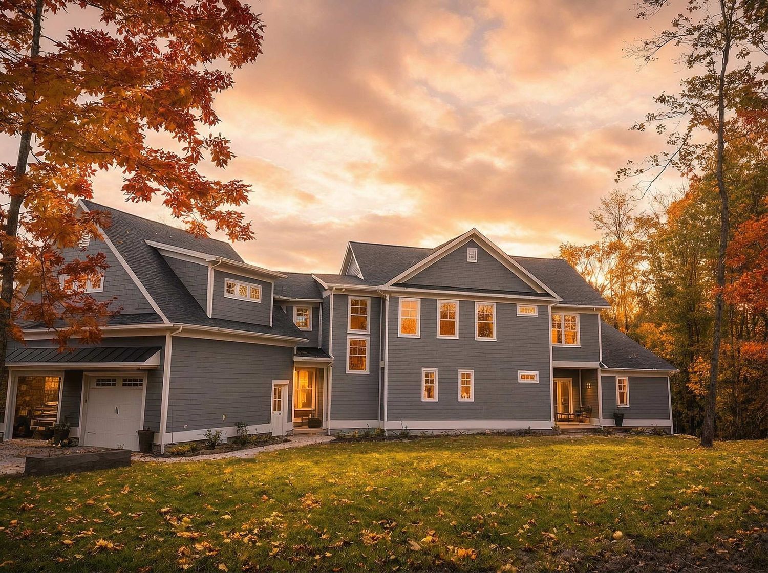Front view of a large gray two story house on wooded slope with covered porch and autumn leaves! around trees.