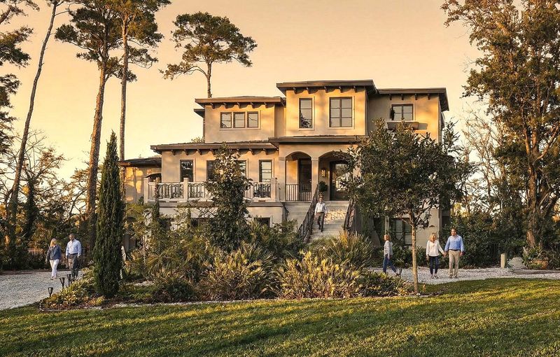 Front view of a large two-story house with arched entryway, light exterior, tall trees, and grassy lawn.